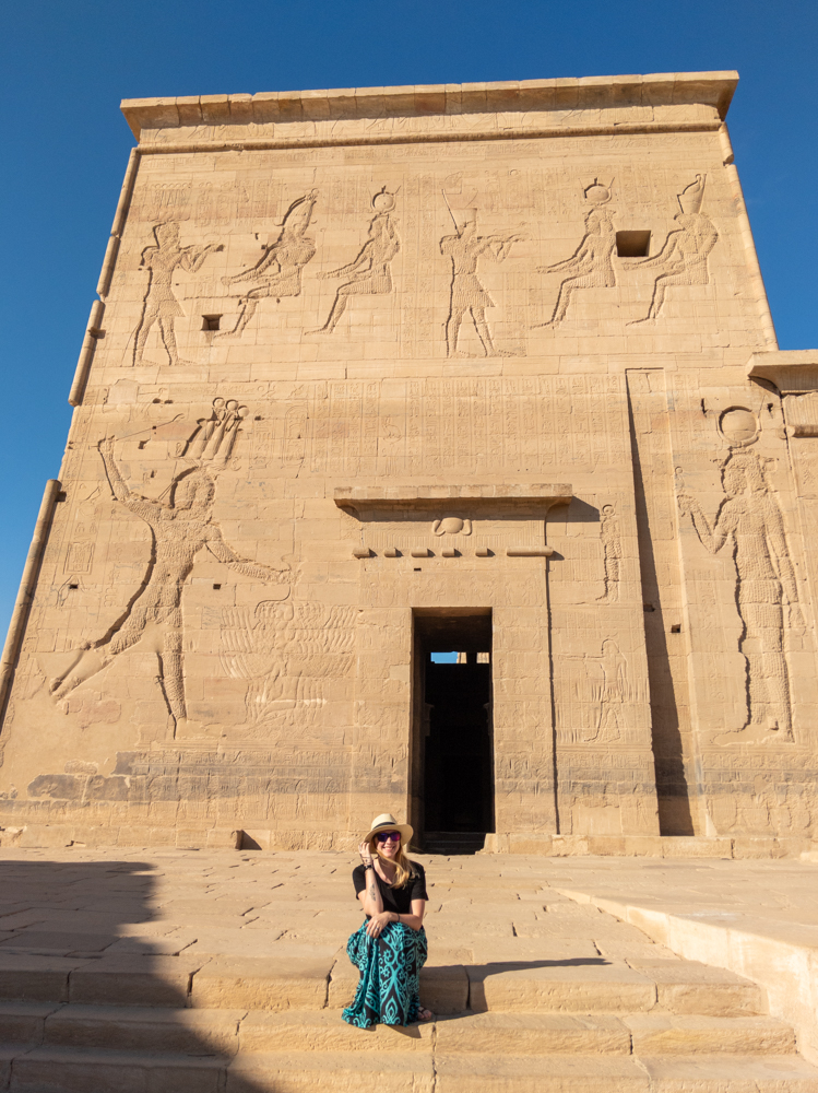 Sitting at Philae Temple in Egypt, wearing long pants and a hat.