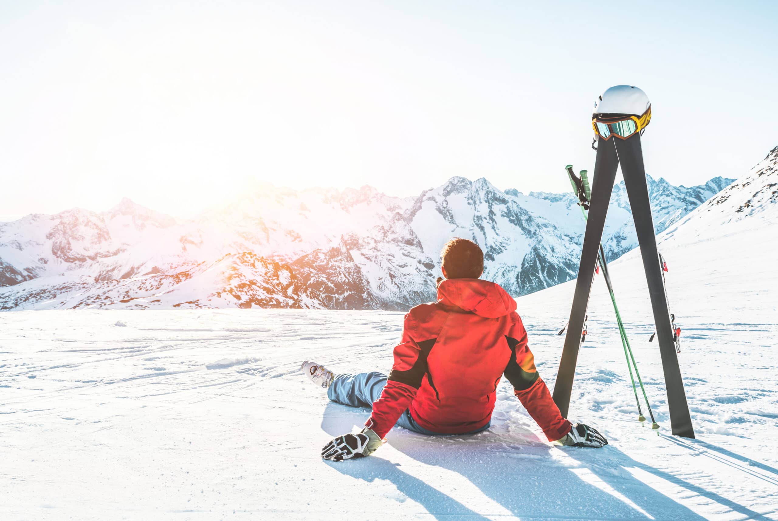 A skier sitting at the top of a mountain with the sun beaming off the distant mountain peaks.