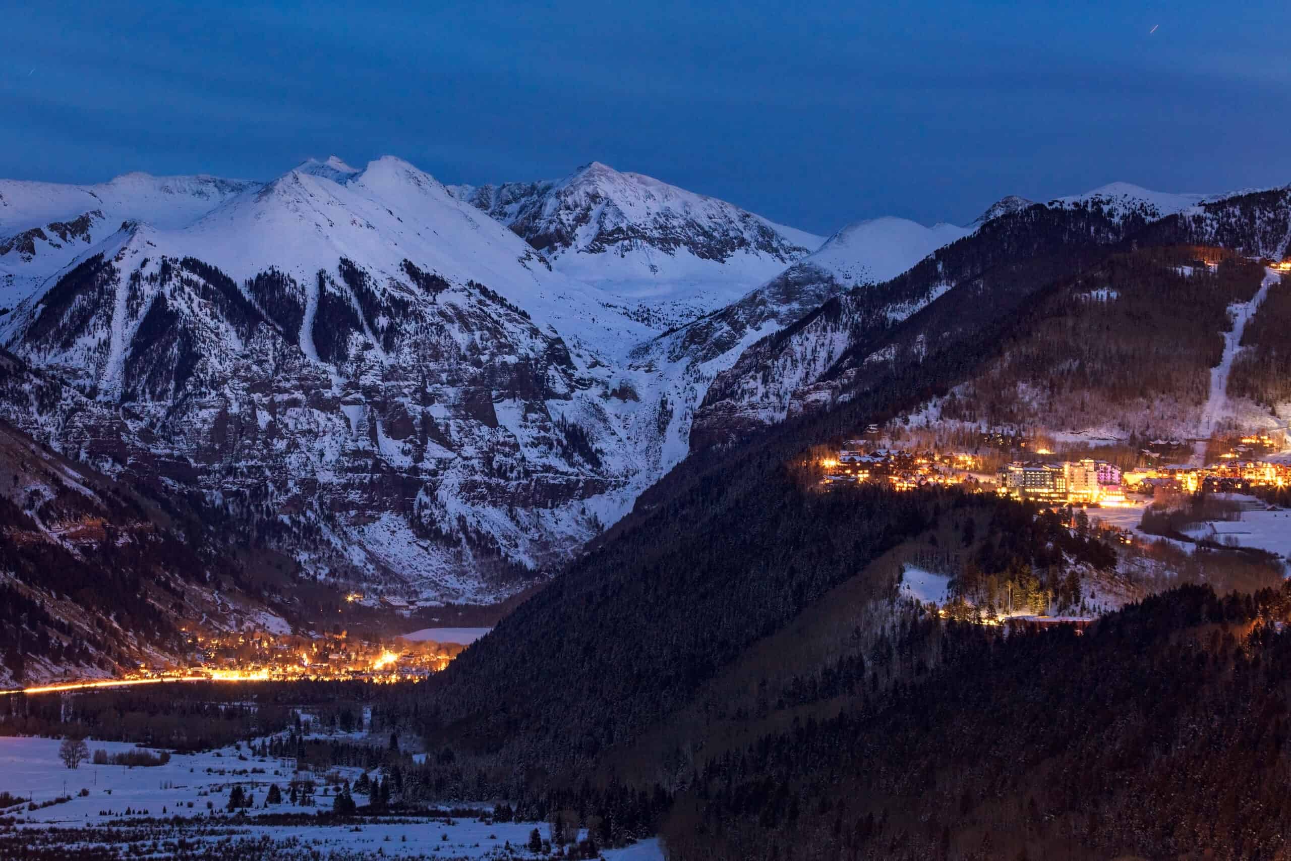 An evening view of Telluride with the town lit up with warm golden lights and the snow mountains towering all around.