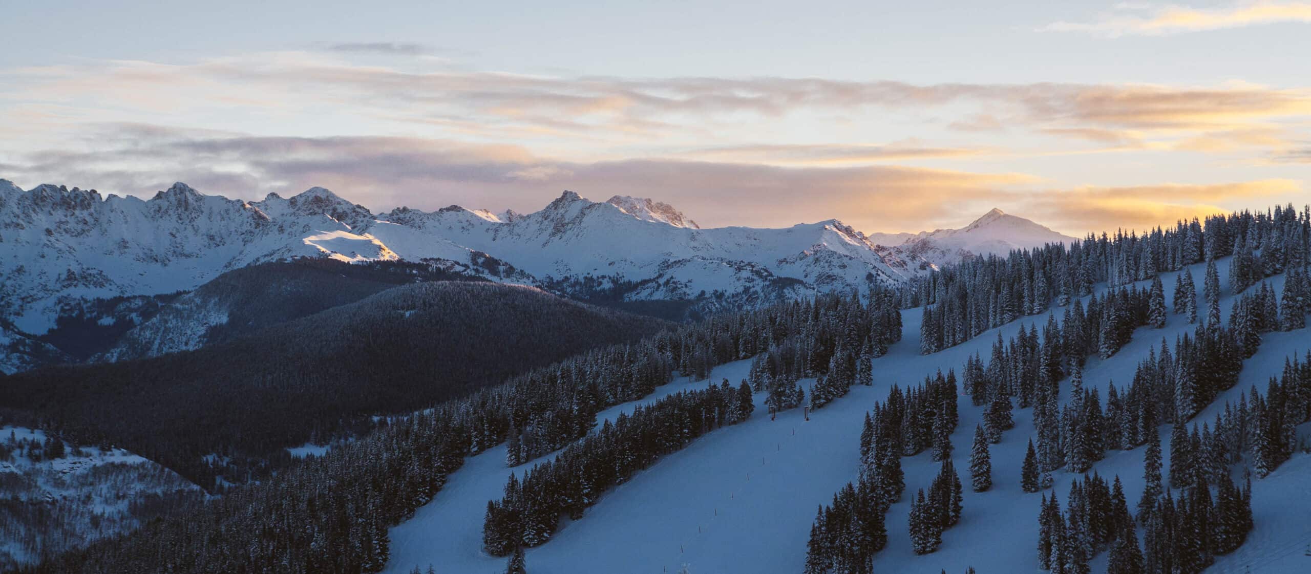 The sun breaks over mountain covered in fresh powder snow and lined with trees.
