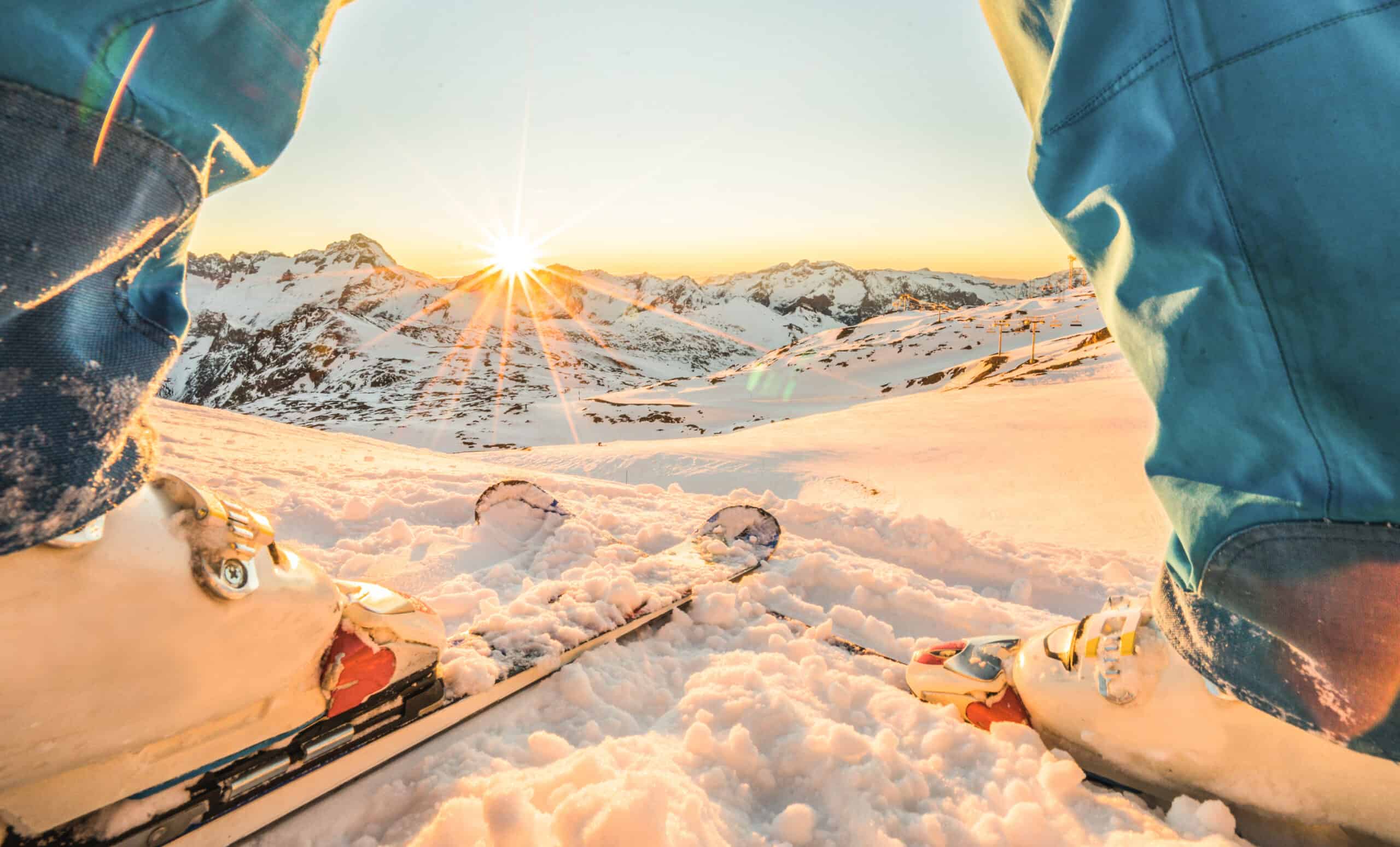 A view through the legs of a skier at the top of a snowy run with the sun peeking over the mountains in the distance.