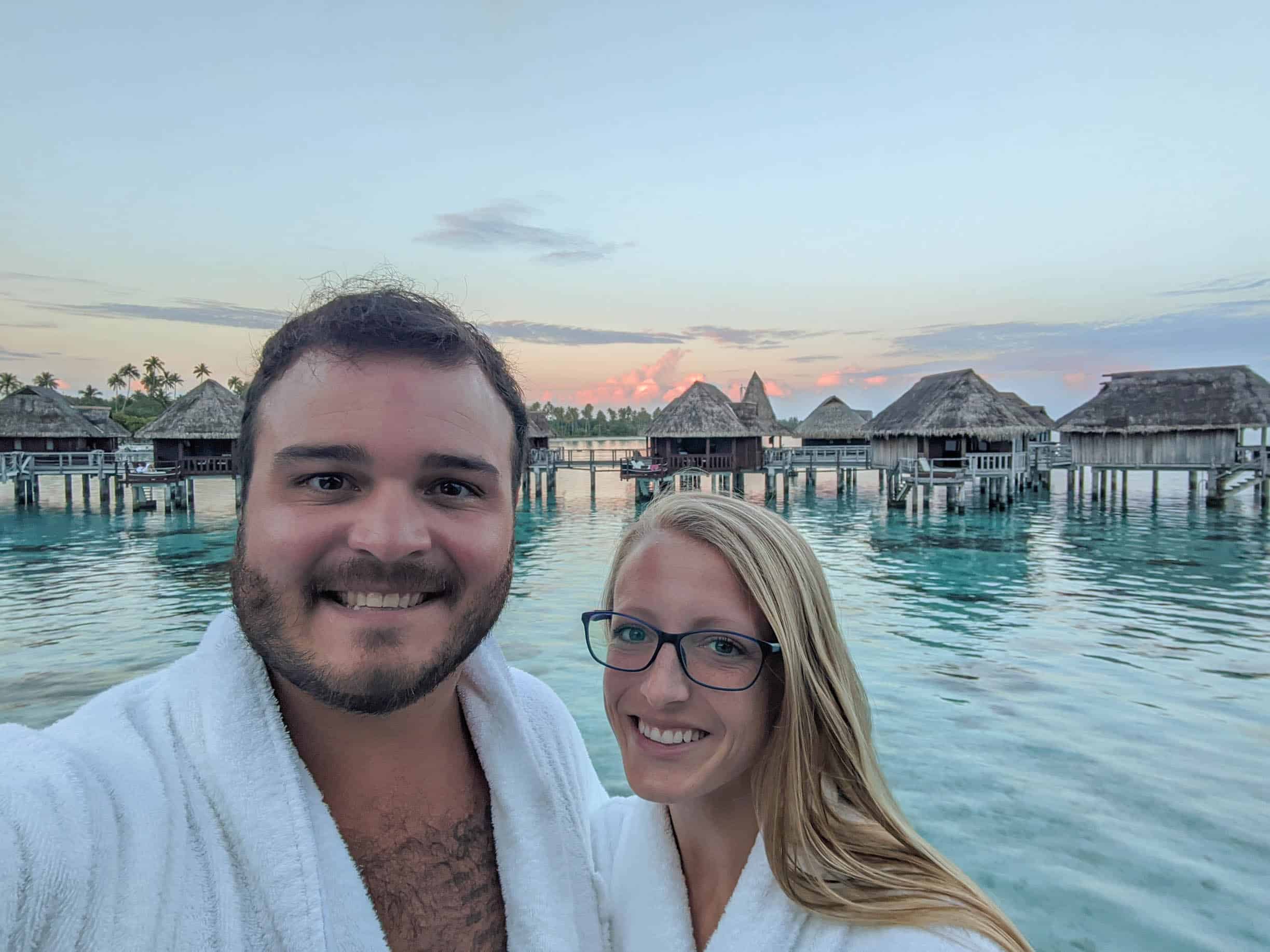 Mike and Laura smiling with the sunsetting behind them with overwater bungalows at the Sofitel Kia Ora Moorea in the distance.