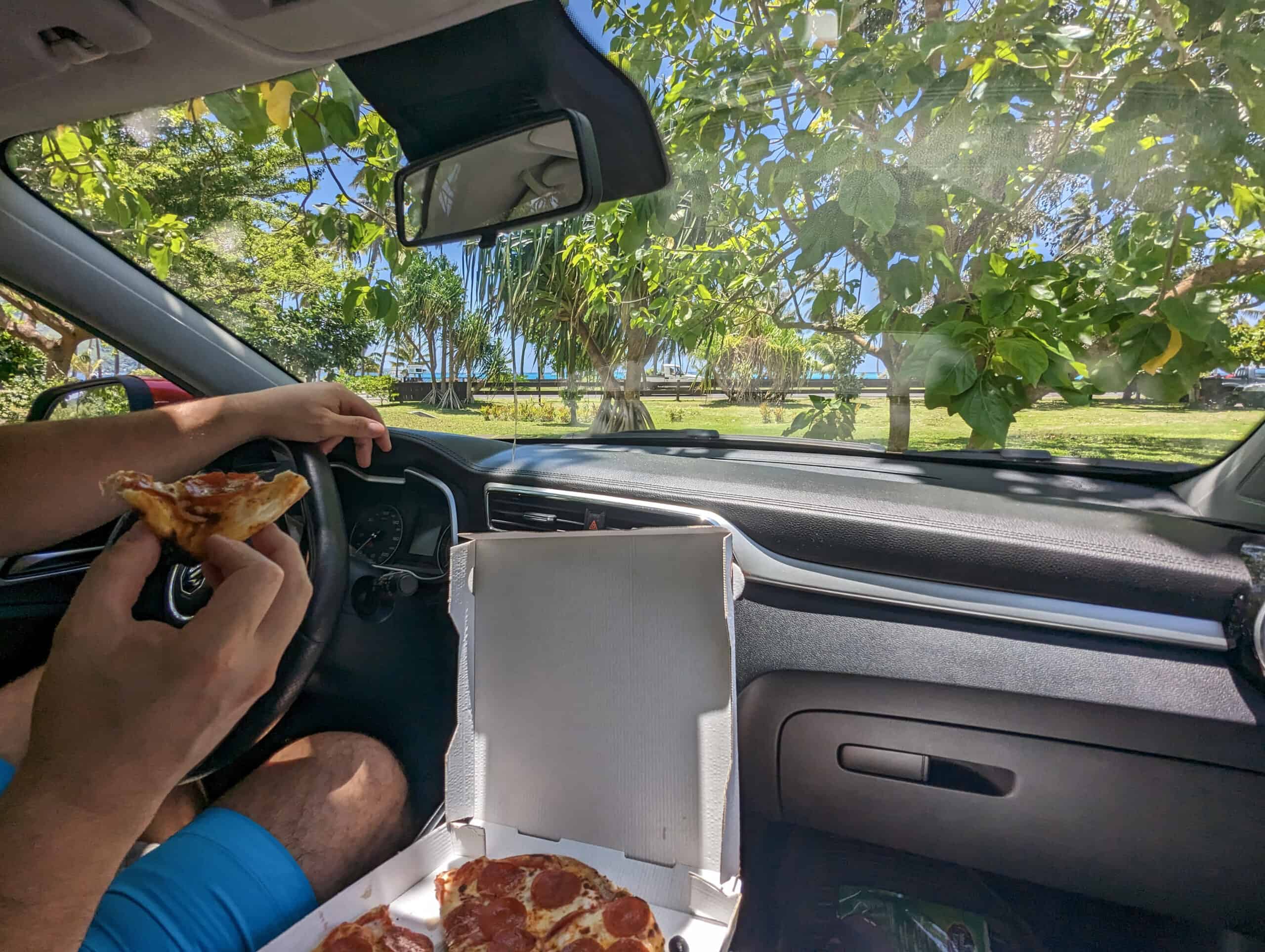 Mike and Laura eat pizza from Allo Pizza as they sit in their electric vehicle rental from Coco Riders in the shade of trees in Moorea.