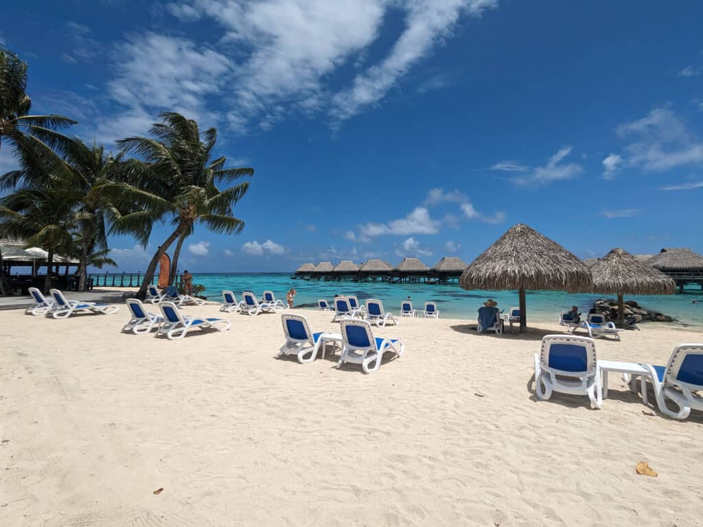 A view of the private beach and umbrellas at the Hilton Moorea with the lagoon and overwater bungalows in the distance.