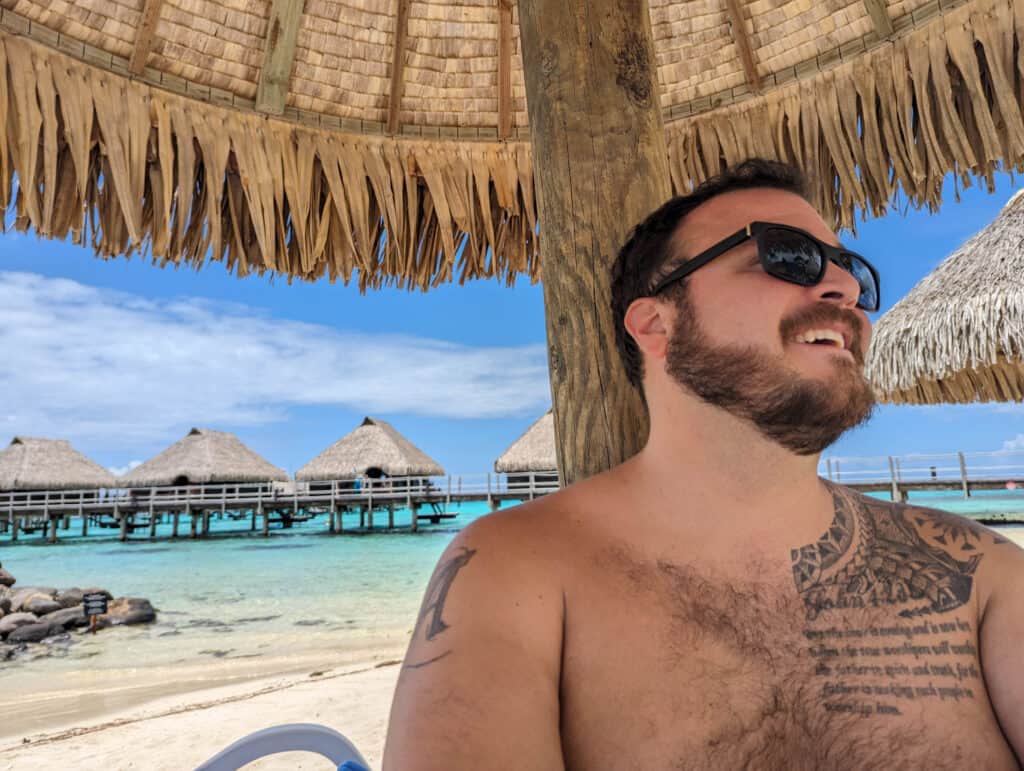 Mike sitting on the beach under an umbrella at the Hilton Moorea with overwater bungalows in the background.