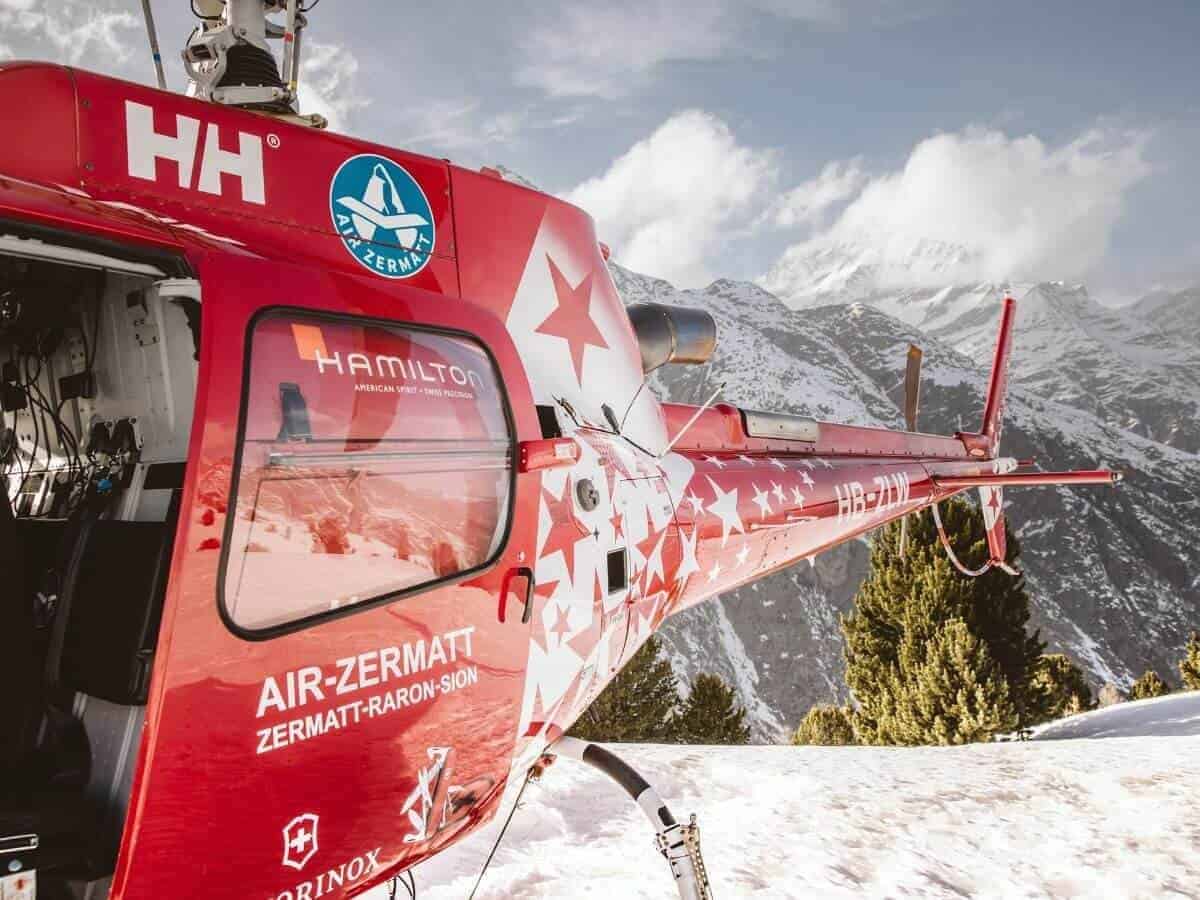 A red Air Zermatt helicopter sits atop a snowy mountain in Zermatt.
