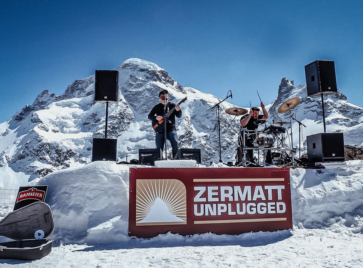 A guitarist and drummer perform on stage at Zermatt Unplugged festival with snowy mountains behind them.